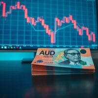 Stack of Australian Dollar banknotes with a blurry background showing a declining AUD/USD chart.