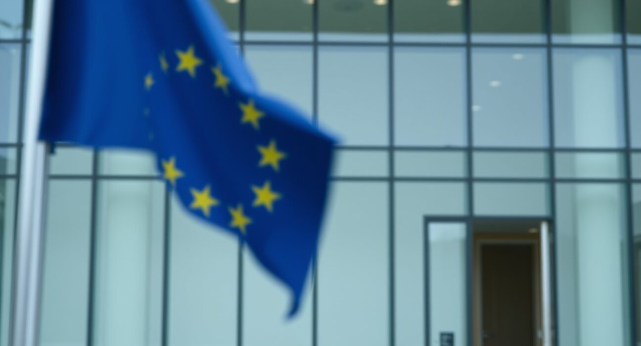 Exterior of a European Union regulatory building with a blurred EU flag in the foreground, representing crypto regulation.