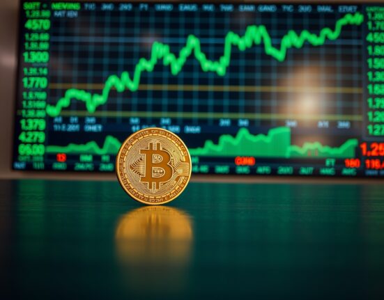 Bitcoin coin on table with stock market display showing upward trends in background
