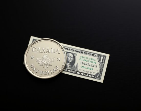 Canadian loonie coin and US dollar bill on a dark surface representing currency market analysis.