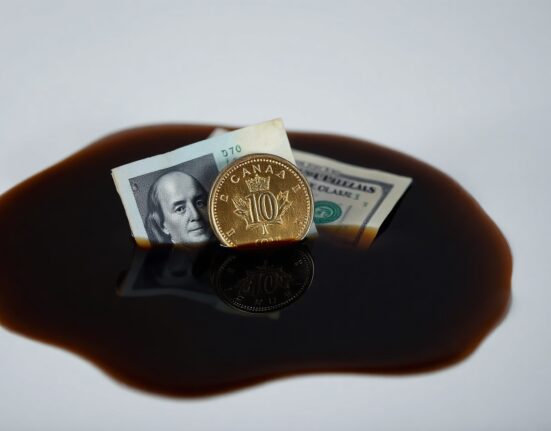Canadian loonie coin and US dollar bill partially submerged in crude oil with a financial chart reflection.