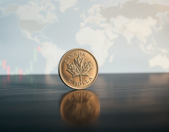 Canadian loonie coin on a wooden surface with a blurred background of financial charts and a world map.