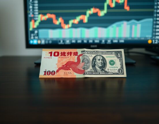 Chinese yuan and US dollar banknotes on a desk with a blurred forex chart in background.