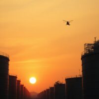 Crude oil storage tanks at sunset with a military helicopter silhouette in the distance.