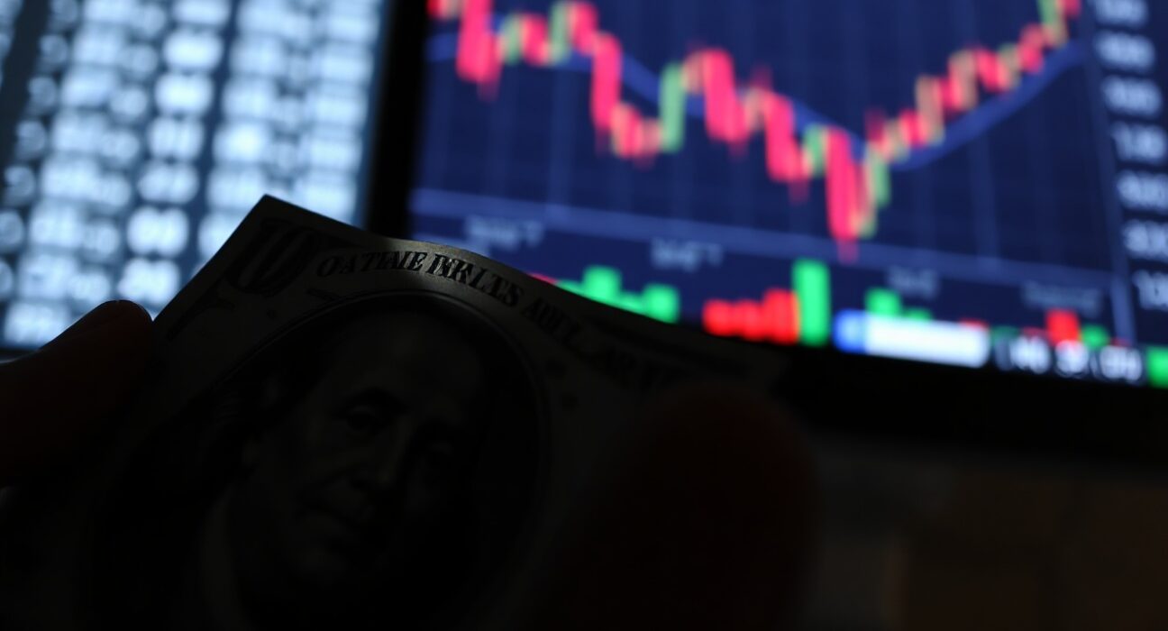 Close-up of a US dollar bill with a downward-trending chart on a trading monitor in the background.