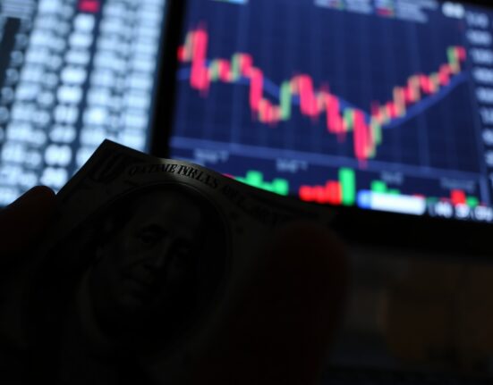 Close-up of a US dollar bill with a downward-trending chart on a trading monitor in the background.