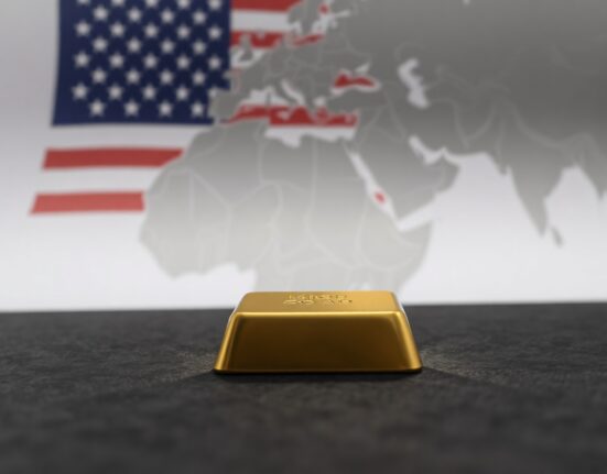Gold bar on dark surface with blurred American flag and Middle East map in background, representing geopolitical market influence.