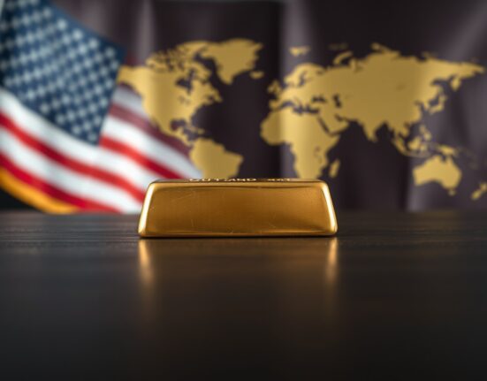 Gold bar on dark wood with US flag and world map in background, representing safe-haven asset and currency market dynamics.