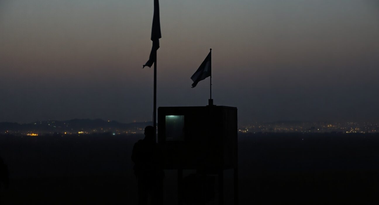 Military checkpoint silhouette at dusk with distant city smoke, representing Iran-Israel tensions
