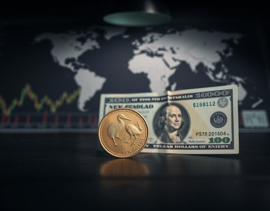 New Zealand Dollar coin and US Dollar bill on a wooden surface with financial charts in background