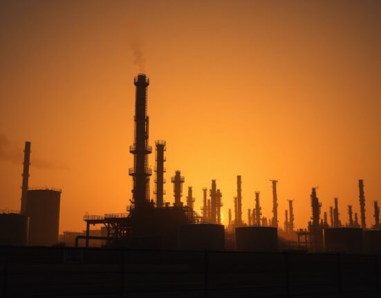 Oil refinery at dusk with industrial towers and flare stacks under a hazy sky, symbolizing geopolitical risk in energy markets.