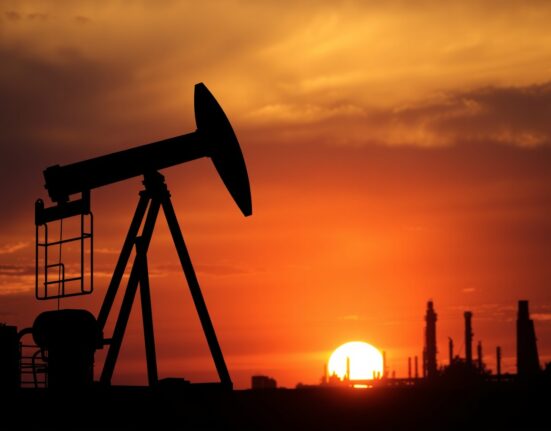 Crude oil pumpjack silhouetted against sunset sky with refinery in background.