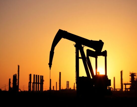 Oil pumpjack silhouetted against sunset with refinery towers in background