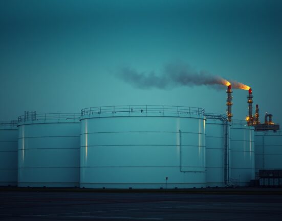 Large white crude oil storage tanks at a refinery under a moody sky, representing oil market volatility.