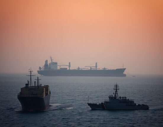Strait of Hormuz at dusk with oil tanker and naval vessel under overcast sky