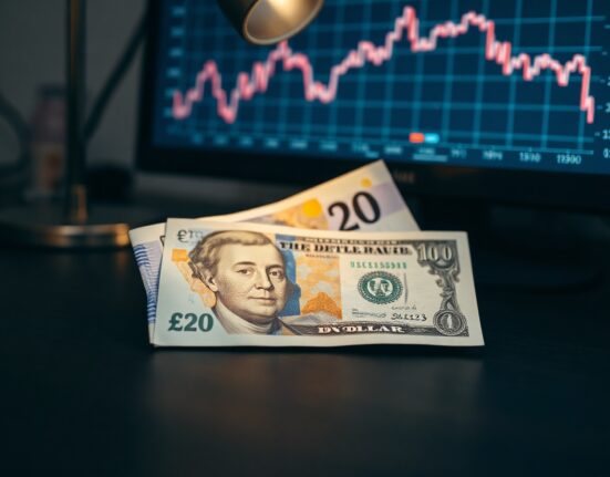 British Pound and US Dollar banknotes on a desk with a computer monitor showing a GBP/USD chart in the background.