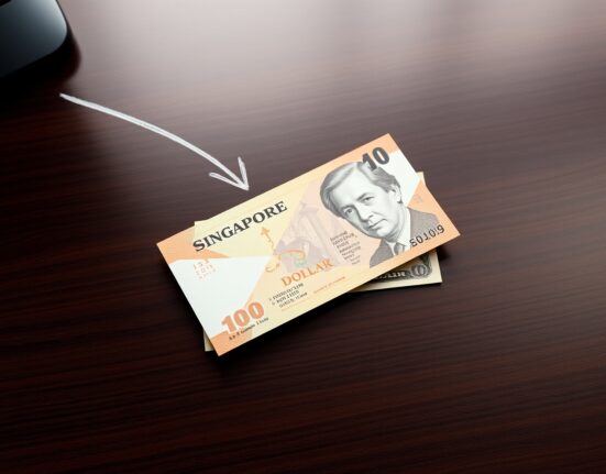 Singapore Dollar banknote with US Dollar bill on desk, downtrend arrow in background.