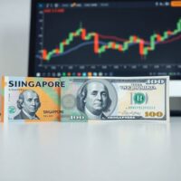 Singapore dollar and US dollar banknotes on a desk with a forex chart in the background.