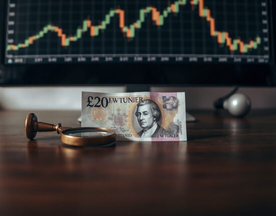 British Pound Sterling note on wooden desk with magnifying glass and blurred financial chart in background