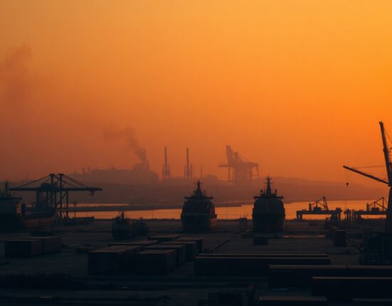 Wide-angle view of a large port with smoke rising from a distant area at dusk
