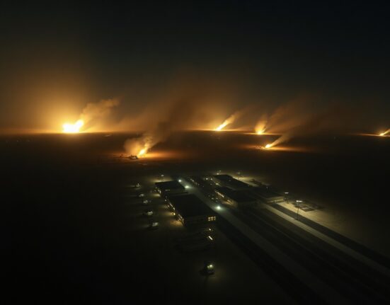 Night aerial view of military installation in desert with explosions and smoke from US airstrikes on Iran