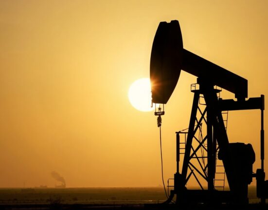 Oil pumpjack silhouetted at sunset with distant smoke, representing geopolitical oil supply risk