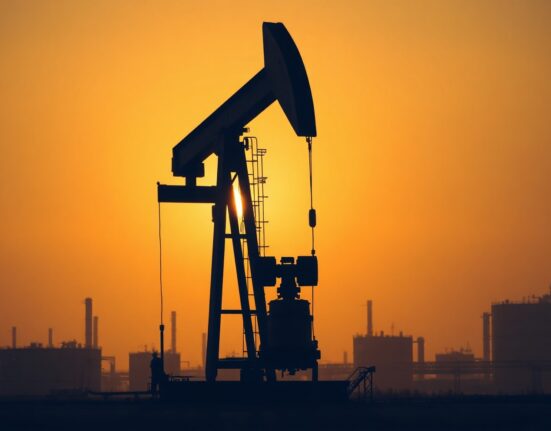 Oil pumpjack at sunset with industrial tanks in background representing crude oil production