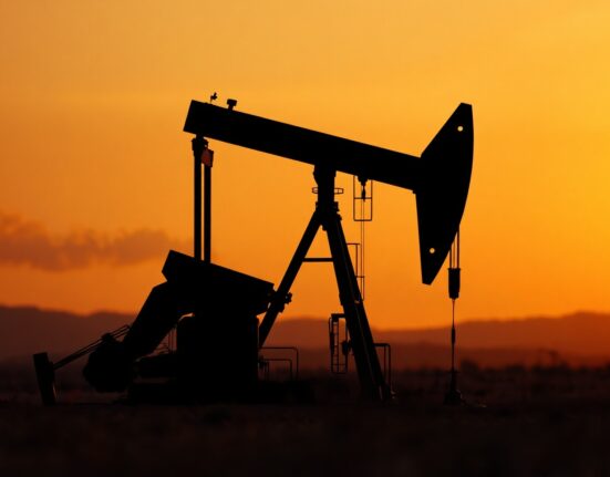 Crude oil pumpjack silhouetted against sunset sky with distant haze, representing geopolitical supply risk