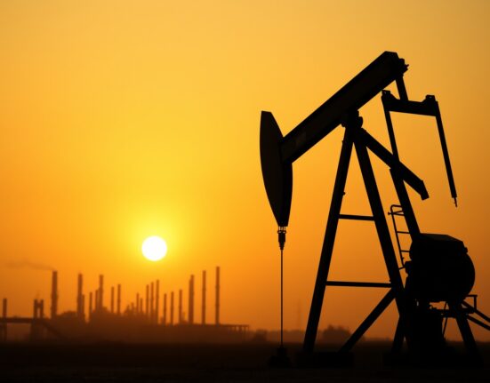 Oil pumpjack silhouetted against a sunset sky near a refinery
