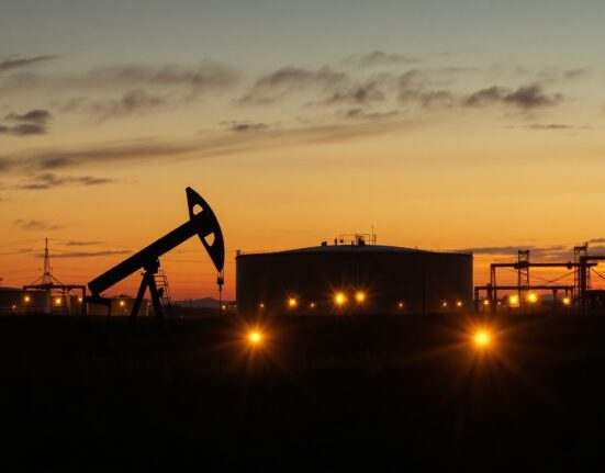 Oil pumpjack silhouette against oil storage tanks at dusk under overcast sky