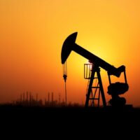 Oil pumpjack silhouetted at sunset with refinery in background representing crude oil supply dynamics