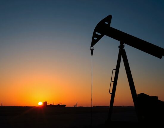 Oil pumpjack silhouetted at sunset with tanker in distance, representing crude supply risks.